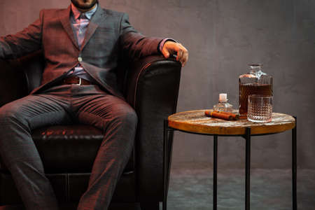 Gentleman sits in a leather armchair with carafe of whisky, empty glass and cigar on the table.の写真素材