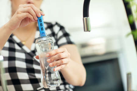 Putting the carbon filter into the bottle. Woman puts carbon filter into the reusable bottle with fresh water in the kitchen. Front view.の写真素材