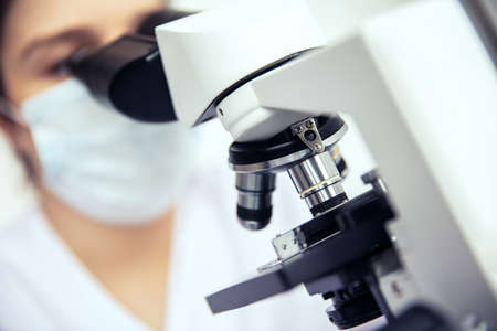 Microscope is used by woman. Medical microscope and female scientist in protective mask in the backgroundの写真素材