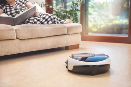 Robot vacuum cleaner working in modern home. Round cleans up living room itself while woman lies on the couch and relaxの写真素材