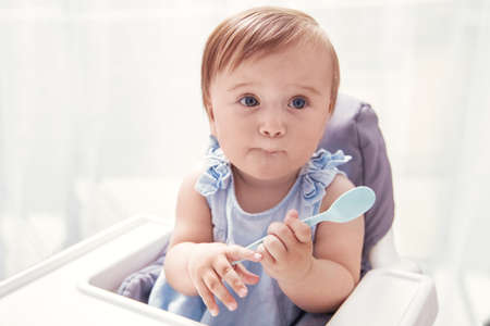 Infant baby girl in blue dress holds baby spoon and makes funny face