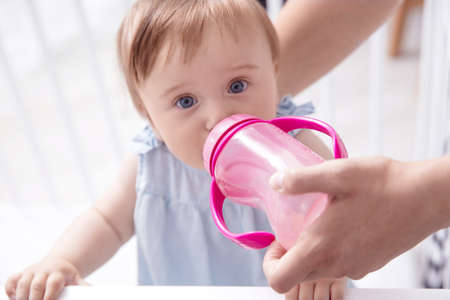 Infant baby girl drinks water from pink sippy cup hold by her mother