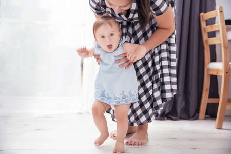 Infant baby girl smiles and learns to walk with assistance of her mother