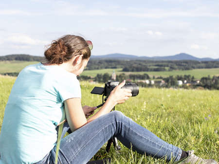 A young young lady takes pictures of the landscape and sits in a meadowの写真素材