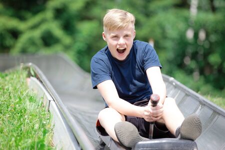 Happy teen boy riding at bobsled roller coaster rail track in summer amusement parkの写真素材