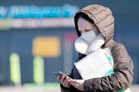 Pandemic virus concept. Woman with medical face mask near supermarket store using the phone to search for internet newsの写真素材
