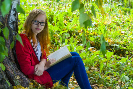 Teenager in glasses and in red jacket sitting in forest and reading a book