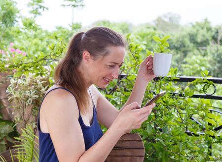 A woman on video call with white cup in hand sitting in a garden and smiling