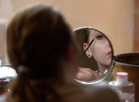 Girl teenager sits in front of window and applies make-up on her eyelashesの写真素材