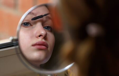 Girl teenager sits in front of round mirror and applies make-up on her eyelashesの写真素材