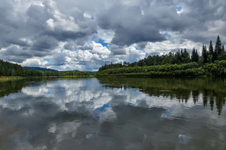 A natural landscape with clouds reflecting on the surface of a calm river with green forest on its...