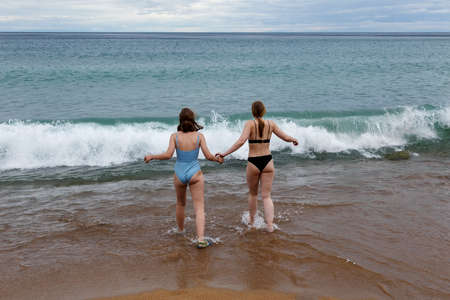 Girls teenagers holding hands in swimming suits on a beach of Baikal lake going to water in cloudy...