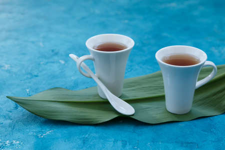 Two white cups with ceramic teaspoon with tea standing on a green leaf with blue backgroundの写真素材