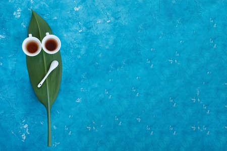 Two white cups with tea standing on a green leaf with blue background, concept face, view from topの写真素材