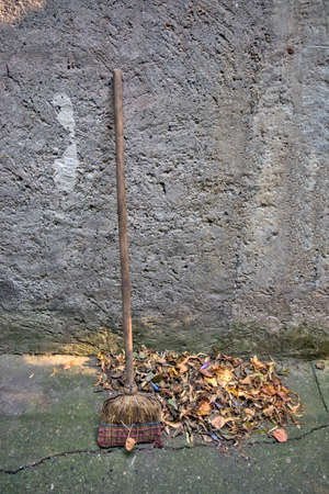 Leaning against the wall stands a worn-out old broom and fallen leaves on a pile.の写真素材