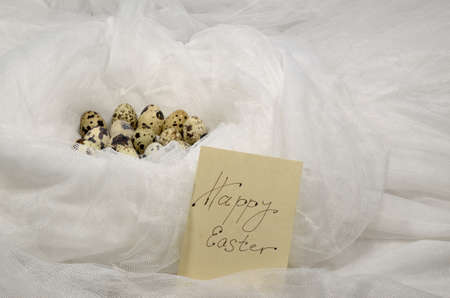 Eggs of quail in white textile nest, with note Happy Easter on a card.の写真素材