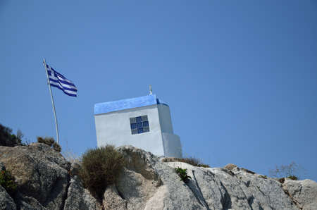 White-blue Greek orthodox chapel on rock with Greek flagの写真素材