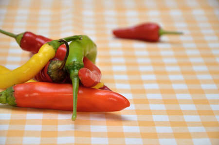 Pile of red, orange, green and yellow hot chili peppers on white-yellow tablecloth with a red pepper...