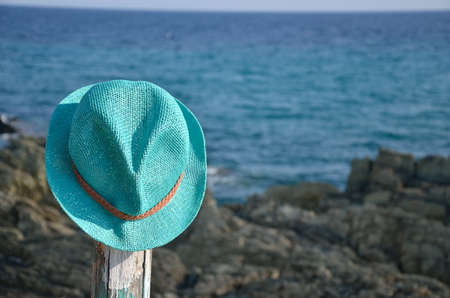 Turquoise straw summer hat left on wooden pillar with rock and sea in backgroundの写真素材