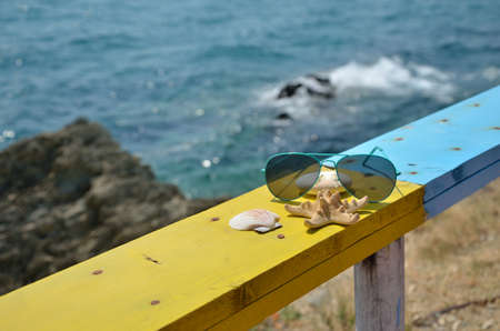 Turquoise sunglasses, sea star, shell and stone on yellow wooden beach bench with turquoise sea in backgroundの写真素材