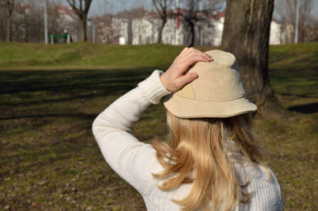 Woman with blond hair in a white sweater holding her hat during a walk in a park on a windy dayの写真素材