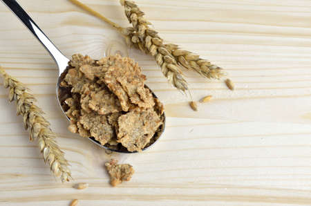 Spoon full of crunchy cereals and wheat ears next to it on wooden table