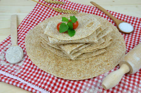 Buckwheat tortillas on red-white tablecloth with cherry tomatoes and fresh leaves on top surrounded...