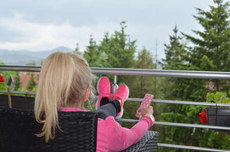 Woman with ponytail in pink sportswear sitting on a balcony and talking over the mobile phone using earbudsの写真素材