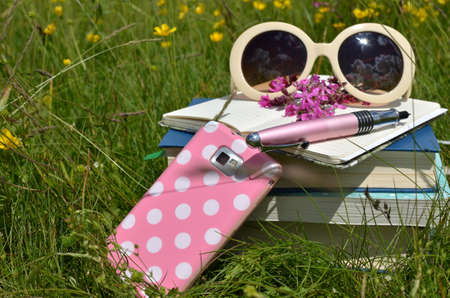 A pile of books, open notebook, wildflowers, pen, elegant sunglasses and mobile phone on green grass...