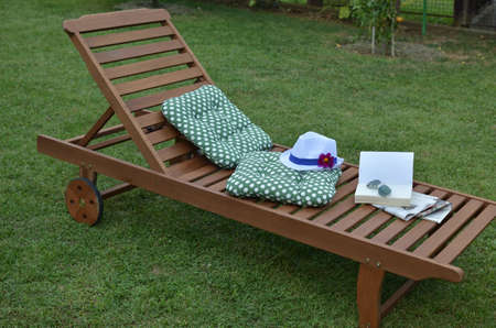 Wooden easy chair on a garden lawn, with cushions, book, sunglasses and hat with flower