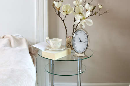 Clock showing eleven and quarter, a book and a cup on a glass table by a bedの写真素材
