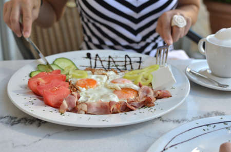 Plate with delicious breakfast - fried eggs, bacon and fresh vegetables - with knife and fork in woman s handsの写真素材