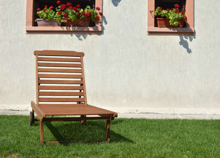 Wooden deck chair on a garden lawn in front of the white house wall with flower potsの写真素材