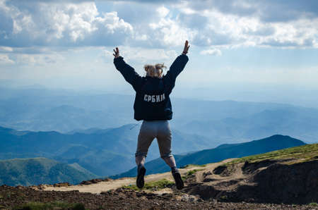 Woman jumping, dressed in a jacked with a Cyrillic title meaning Serbia, watching idyllic landscape of the mountain Kopaonik, in Serbia, in summerの写真素材