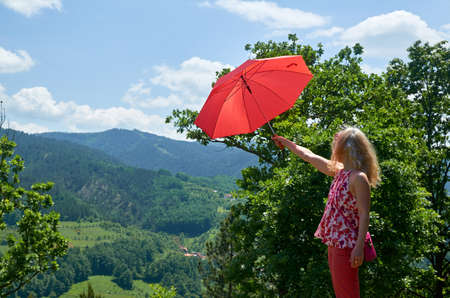 Woman dressed in red holding red umbrella used as a parasol while enjoying a picturesque view of a landscape on a sunny dayの写真素材