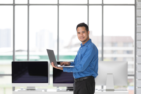 Young Asian businessman standing and holding new laptop computer with happiness manner in modern office, group of desktop computers blur in beside.の写真素材