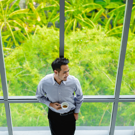 Young businessman holding a cup of coffee standing by the window looking away, with a green tree as...