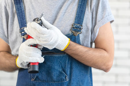 Young Asian man holding power drill standing in front of white brick wall, smiling and looking at camera, concept for home DIYの写真素材