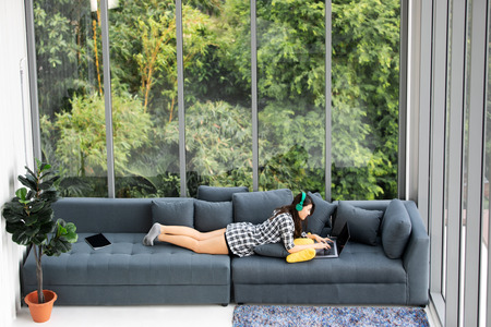 Asian woman laying on sofa near big glass wondows, relaxing alone in house with green forest in backgroundの写真素材