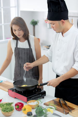 Asian male chef demonstrating how to cook for beautiful student with smoke from black pan in white clean modern kitchen.の写真素材