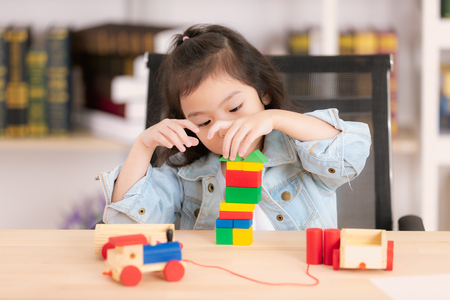 Lovely cute little Asian girl in jeans shirt playing wood block toys on desk. Concept for funny activity of young kids in free time.の写真素材