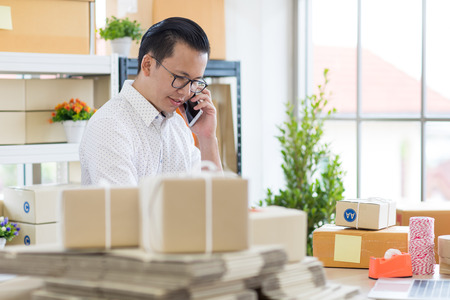 Young Asian businessman in casual shirt use calling smartphone,  working in simple house office look like doing startup business. Concept for online marketing, SME and home base workplace.の写真素材
