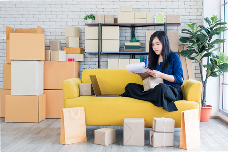 Young and beautiful Asian woman sitting among several boxes and checking parcels on the yellow sofa, working in the house office. Concept for home base business and startup ownership.の写真素材