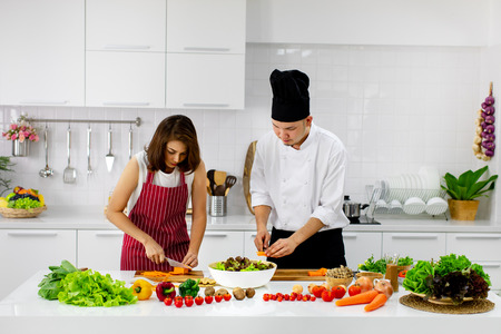Beautiful Asian woman in red apron learning how to cook and mix vegetable salad in big bowl, teaching by Asian chef in white modern kitchen.の写真素材