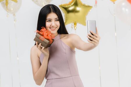Attractive young asian woman in pink dress, selfie with her gift box at fun party, white background with festive colorful balloons.の写真素材