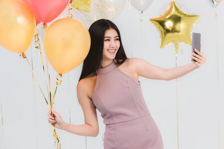 Attractive young asian woman in pink dress, selfie with colorful balloons at fun party, portrait shot on white background.の写真素材