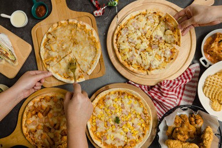 Top view shot of various kinds of delicious tasty juicy cheesy pizza placed on wooden board on party table around with other fast food while unrecognizable people picking up eating dinner together.の写真素材