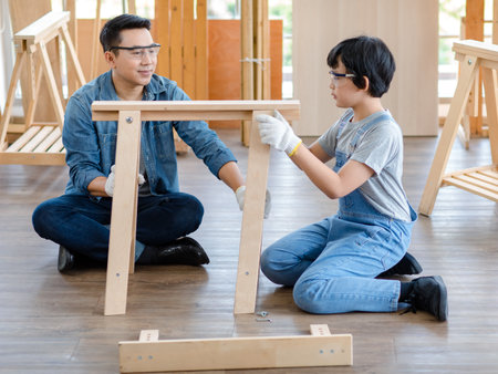 Asian amateur young boy in jeans outfit with safety glasses goggles and gloves helping professional carpenter engineer father holding assembling wooden furniture in housing building construction site.の写真素材