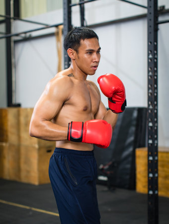 Asian young muscular fit strong body sporty athletic shirtless male fitness model in red boxing gloves with tattoo standing guarding footwork workout exercising sparring training alone in Crossfit gymの写真素材