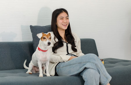 Asian young happy cheerful female owner sitting smiling on cozy sofa couch holding eating popcorn glass bowl snack watching movie with best friend companion dog white short hair jack russell terrier.の写真素材